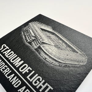 Puede incluir: Un posavasos cuadrado de pizarra negra con un grabado l&aacute;ser blanco detallado de una vista a&eacute;rea del Stadium of Light. El texto "STADIUM OF LIGHT" y "SUNDERLAND AFC" est&aacute; impreso en una fuente blanca sans-serif en la parte inferior izquierda.