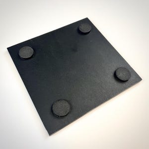 Puede incluir: Un posavasos cuadrado de color negro con una superficie lisa y plana, que cuenta con cuatro pies circulares de goma negra fijados en las esquinas inferiores para proporcionar estabilidad y evitar que se deslice al usarlo.