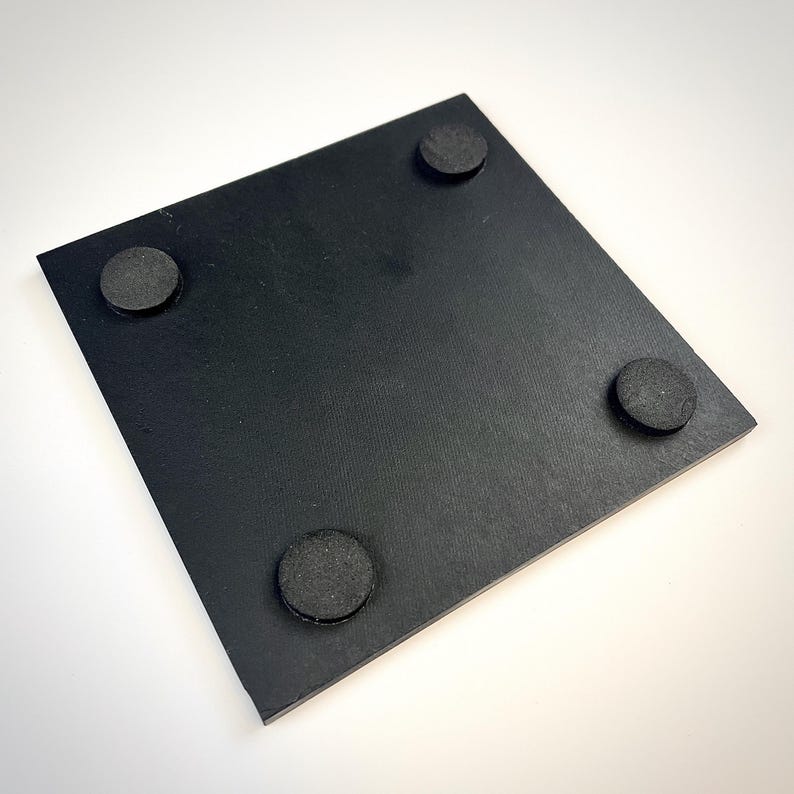 Puede incluir: Un posavasos cuadrado de color negro con una superficie lisa y plana, que cuenta con cuatro pies circulares de goma negra fijados en las esquinas inferiores para proporcionar estabilidad y evitar que se deslice al usarlo.