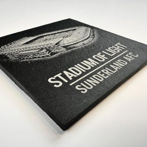 Puede incluir: Un posavasos cuadrado de pizarra negra con una ilustraci&oacute;n grabada con l&aacute;ser en blanco del Stadium of Light. El texto STADIUM OF LIGHT y SUNDERLAND AFC est&aacute; impreso en letras may&uacute;sculas blancas debajo del gr&aacute;fico arquitect&oacute;nico del estadio.