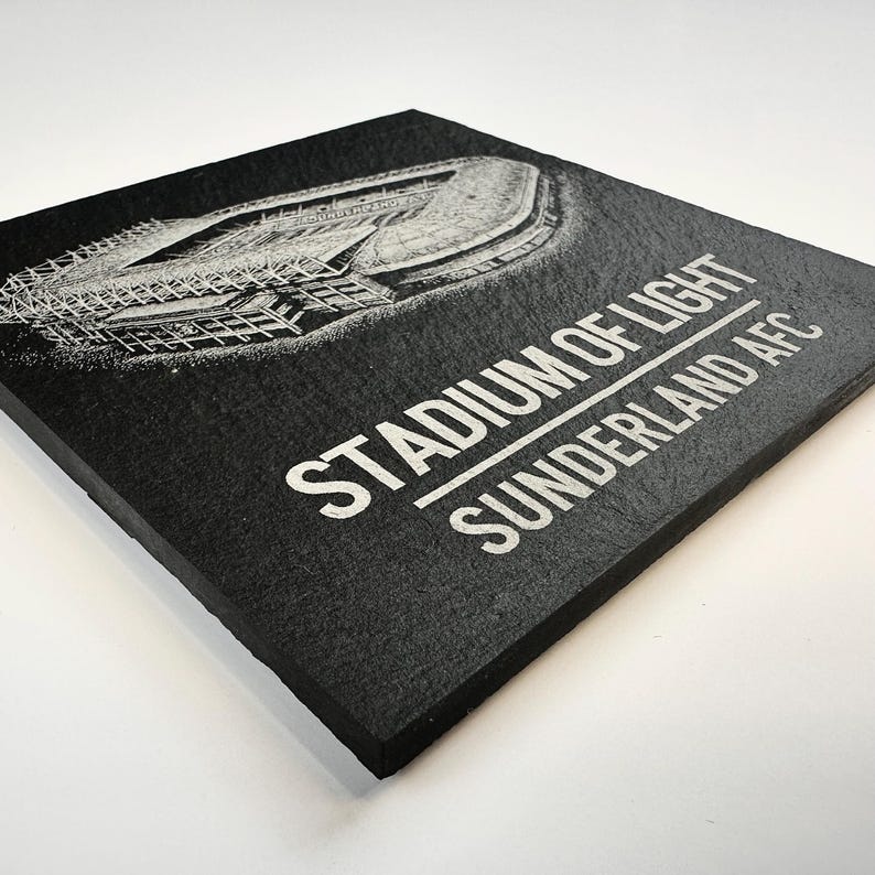 Puede incluir: Un posavasos cuadrado de pizarra negra con una ilustraci&oacute;n grabada con l&aacute;ser en blanco del Stadium of Light. El texto STADIUM OF LIGHT y SUNDERLAND AFC est&aacute; impreso en letras may&uacute;sculas blancas debajo del gr&aacute;fico arquitect&oacute;nico del estadio.