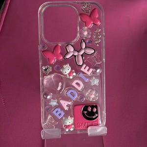 Può includere: Una custodia per telefono trasparente con un design rosa, viola e bianco. La custodia presenta un personaggio Hello Kitty, un'emoticon sorridente, farfalle e la parola "BADDIE" su di essa.