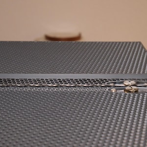 May include: A close-up of a black metal box with a honeycomb pattern on the top and bottom. The box has a hinge on the side and a silver latch.