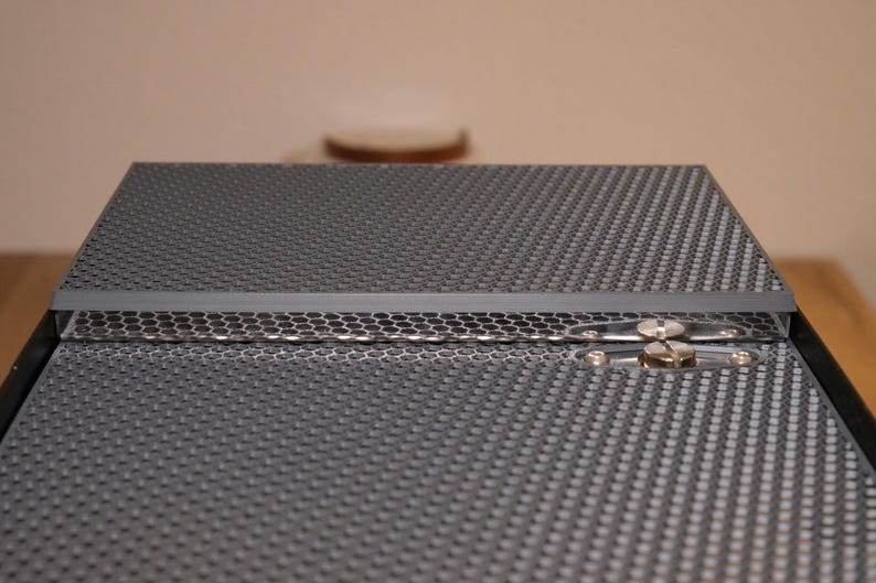 May include: A close-up of a black metal box with a honeycomb pattern on the top and bottom. The box has a hinge on the side and a silver latch.