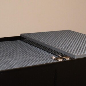 May include: A black box with a gray textured lid. The lid is slightly ajar, revealing the interior of the box, which is also lined with a gray textured material.