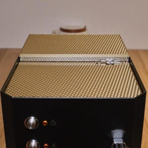 May include: A black espresso machine with a gold-colored top and a textured surface. The machine has two knobs and a small light on the front.