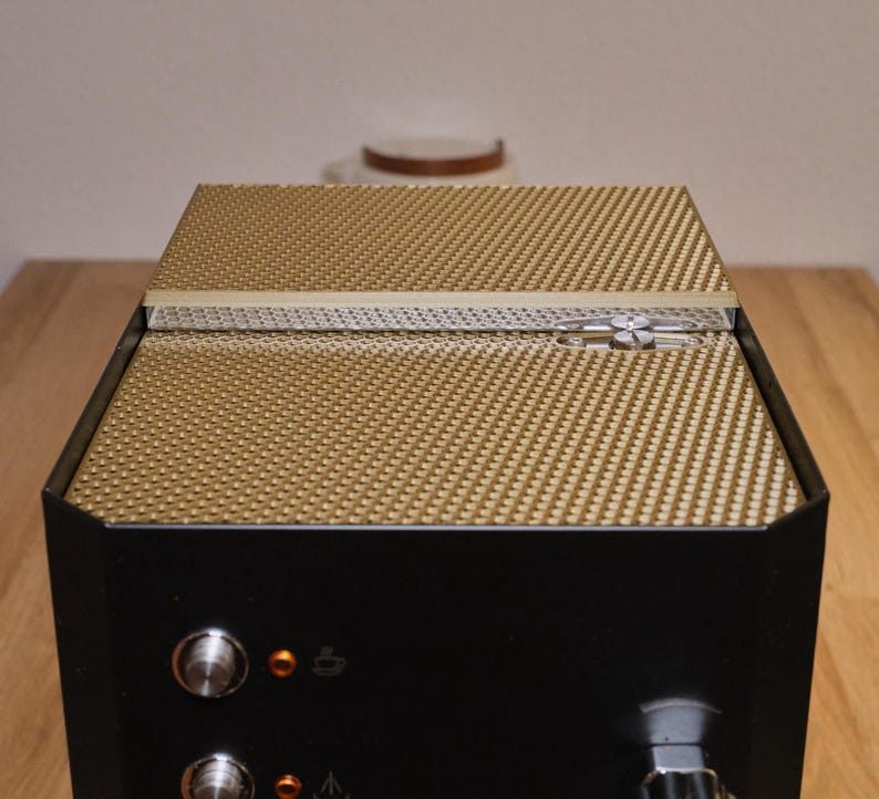 May include: A black espresso machine with a gold-colored top and a textured surface. The machine has two knobs and a small light on the front.