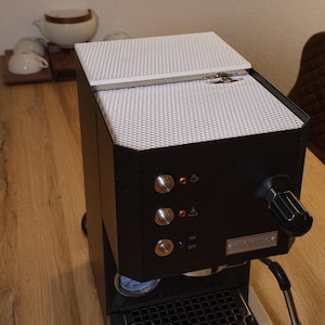 May include: A black espresso machine with a white lid and a silver drip tray. The machine has a pressure gauge, a steam wand, and a control panel with buttons for on/off, steam, and hot water.