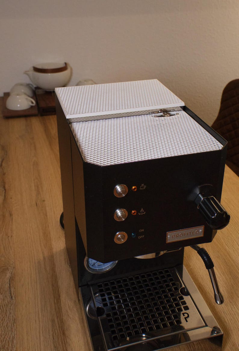 May include: A black espresso machine with a white lid and a silver drip tray. The machine has a pressure gauge, a steam wand, and a control panel with buttons for on/off, steam, and hot water.