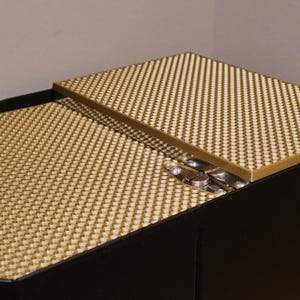 May include: A black box with a gold-colored lid. The lid is made of a textured material and has a hinge. The box is closed.