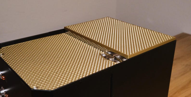 May include: A black box with a gold-colored lid. The lid is made of a textured material and has a hinge. The box is closed.