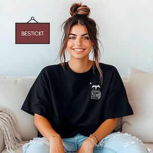 Könnte beinhalten: Eine Frau trägt ein schwarzes T-Shirt mit einer weißen Grafik einer Fledermaus, die über einem Stapel Bücher fliegt. Das Shirt hat das Wort "BESTICKT" auf einem Etikett aufgedruckt.