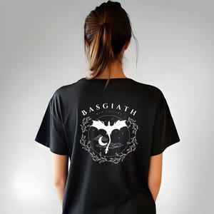 Könnte beinhalten: Schwarzes T-Shirt mit einem weißen Aufdruck eines Drachen, der mit einer zunehmenden Mondsichel und Sternen fliegt. Der Text "BASGIATH WAR COLLEGE" befindet sich über dem Drachen.