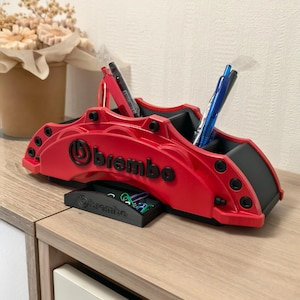 Peut inclure: Un organisateur de bureau rouge et noir en forme d'étrier de frein Brembo avec plusieurs compartiments pour stylos et un petit plateau pour trombones. Le mot Brembo est imprimé en noir sur le côté du corps principal et sur le devant du plateau.