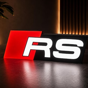 May include: A rectangular illuminated sign featuring the letters RS in bold white font. The left side of the sign displays a solid red square background, while the right side features the white letters RS set against a black background.
