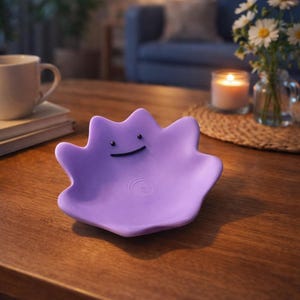 Peut inclure: Un plat violet en forme de personnage souriant. Il est posé sur une table en bois, avec une tasse, des livres, des fleurs et une bougie en arrière-plan. Le plat présente un motif circulaire au centre.