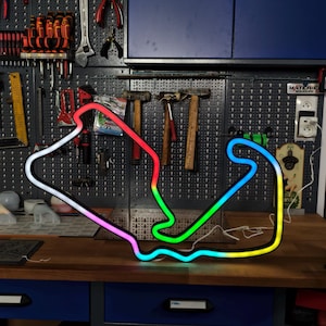 Peut inclure: Une enseigne néon représentant un circuit de course de Formule 1. La lumière présente un dégradé de couleurs vives passant du rouge et blanc au vert, bleu et jaune le long de son tracé sinueux et continu sur un fond sombre.