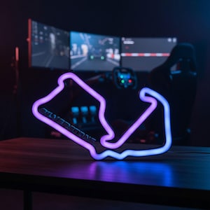 May include: A neon sign shaped like the Silverstone race track circuit, featuring a gradient of purple and blue light. The word Silverstone is embossed in dark lettering along the lower left section of the track outline, glowing against the dark.