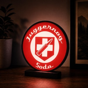Peut inclure: Une enseigne lumineuse circulaire avec un fond rouge, une croix blanche et un graphique de balle au centre. Le texte Juggernog est inscrit en arc de cercle en haut, et le mot Soda est imprimé en bas dans une police blanche stylisée.