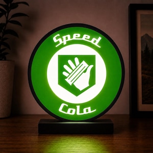 May include: A circular LED sign featuring a bright green background with a white border and a central white icon of a hand. The text Speed Cola is printed in white lettering at the top and bottom of the circle. The sign sits on a black base.
