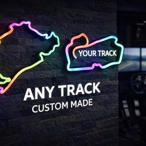Puede incluir: Letreros de neón con forma de circuitos de carreras, con el texto "ANY TRACK CUSTOM MADE" y "YOUR TRACK". Los letreros tienen un efecto degradado colorido y están montados en una pared oscura y texturizada.