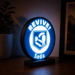 May include: A circular, illuminated sign featuring a blue and white design with the text "REVIVE!" at the top and "Soda" at the bottom. The centre displays a white shield icon containing a stylised graphic of a person standing by a tombstone.