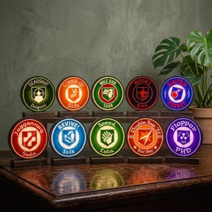Könnte beinhalten: Zehn kreisförmige Leuchtschilder mit verschiedenen grafischen Logos und Texten wie Deadshot Daiquiri, Stamin-Up, Mule Kick, Widow's Wine, Electric Cherry, Juggernog, Revive, Speed Cola, Double Tap II und Flopper PHD, jeweils auf einem Holzsockel.