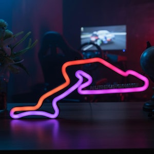 Peut inclure: Une enseigne néon en forme du circuit de course Nürburgring, présentant un dégradé de lumière orange et violette. Le mot Nürburgring est clairement visible en lettres sombres intégrées dans la partie inférieure du tracé lumineux.