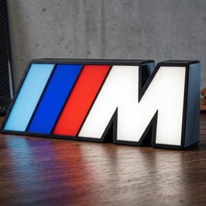 Pode incluir: Um letreiro retangular iluminado com a letra M em branco, precedida por três faixas inclinadas em azul claro, azul escuro e vermelho. O letreiro tem uma moldura preta e foi concebido para ficar na vertical sobre uma superfície plana.