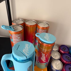 May include: Several aluminum cans of Alani Nu energy drinks, some with blue insulated holders. The cans are orange and pink with the words "ORANGE KISS" and "ENERGY" printed on them. Some cans have the Nutrition Facts label visible.
