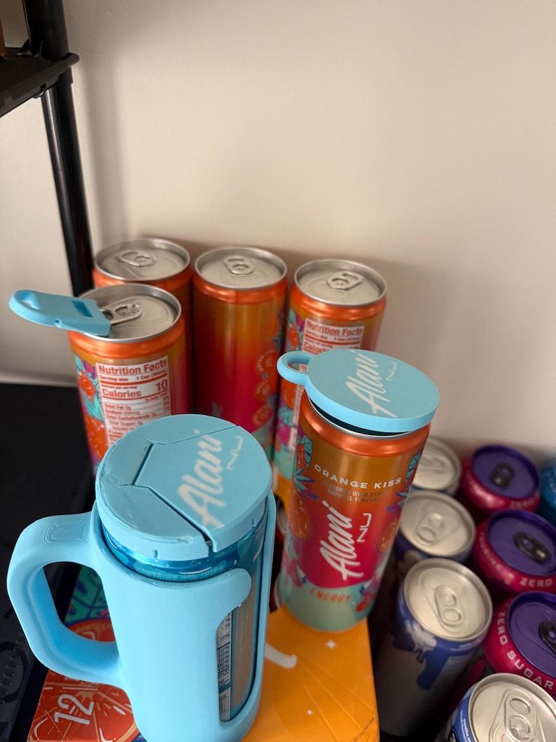 May include: Several aluminum cans of Alani Nu energy drinks, some with blue insulated holders. The cans are orange and pink with the words "ORANGE KISS" and "ENERGY" printed on them. Some cans have the Nutrition Facts label visible.