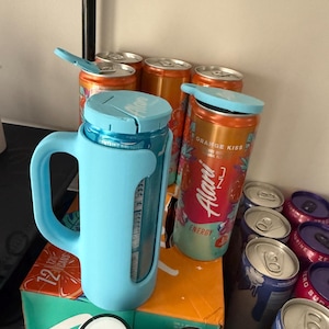 May include: A light blue insulated mug with a handle, holding a can of orange-flavored energy drink. Several other cans of energy drinks are visible, some with the lids open. The cans are orange and blue, with the brand name "Alani Nu" visible.