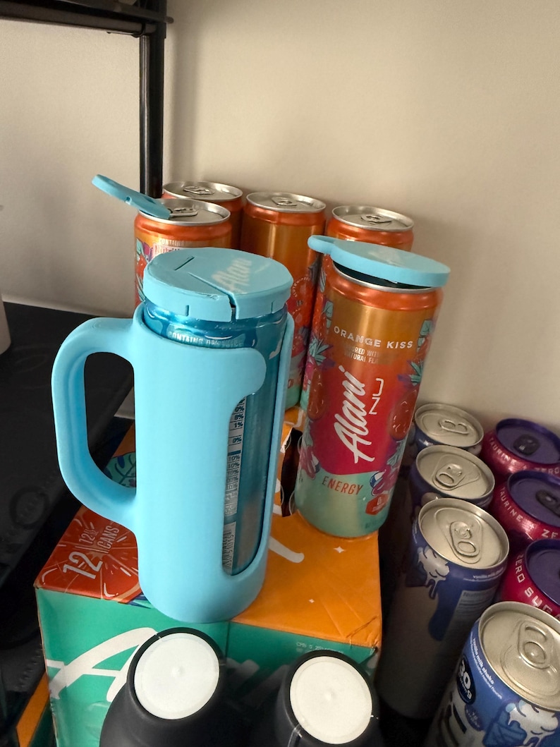 May include: A light blue insulated mug with a handle, holding a can of orange-flavored energy drink. Several other cans of energy drinks are visible, some with the lids open. The cans are orange and blue, with the brand name "Alani Nu" visible.
