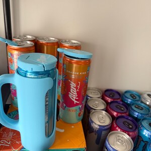May include: A collection of energy drink cans in various colors, including orange, blue, and purple. Some cans are in a light blue holder with a handle. The cans have the text "Alani Nu Energy" and "Orange Kiss".