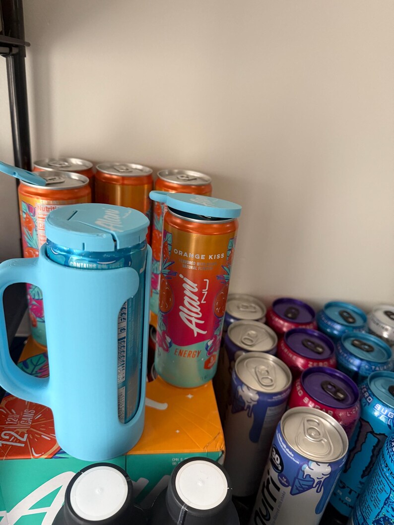 May include: A collection of energy drink cans in various colors, including orange, blue, and purple. Some cans are in a light blue holder with a handle. The cans have the text "Alani Nu Energy" and "Orange Kiss".