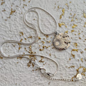 May include: Silver-toned necklace featuring a round pendant with a sun, moon, and star design. The pendant is intricately detailed and hangs from a delicate chain with a lobster clasp. The necklace is displayed on a textured white surface.