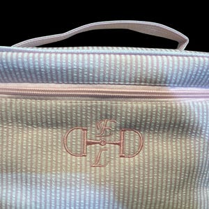 May include: A pink and white striped cosmetic bag with a pink zipper and handle. The bag features an embroidered monogram in pink thread. The bag is designed to hold makeup and other beauty products.