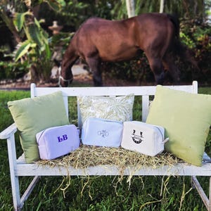 May include: Three striped cosmetic bags with embroidered initials and a smiling face design rest on a white bench with green pillows. A brown horse grazes in the background, creating a serene outdoor scene.