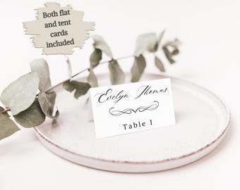May include: A white place card with the name "Evelyn Thomas" in elegant script and the text "Table 1". The card is on a white plate, with a sprig of greenery. Text on a sign says "Both flat and tent cards included".