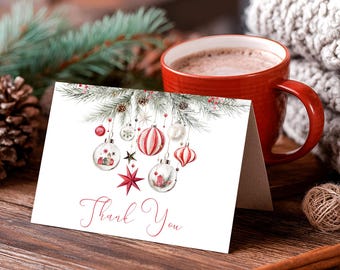 May include: A folded white greeting card with the words "Thank You" in red script. The card features watercolor illustrations of Christmas ornaments and pine branches. A red mug of coffee sits next to the card on a wooden surface.