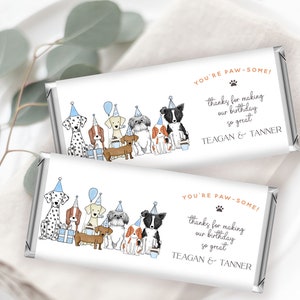 Puede incluir: Dos envoltorios de barra de chocolate blanco con un tema de fiesta de perros azul y blanco. Los envoltorios presentan una línea de perros con sombreros de fiesta y sosteniendo regalos. El texto en los envoltorios dice "You're Paw-some!" y "Gracias por hacer que nuestro cumpleaños sea tan genial." Los nombres "Teagan & Tanner" están impresos en la parte inferior de los envoltorios.