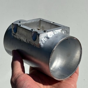 Könnte beinhalten: Ein silbernes Aluminiumrohr mit einer rechteckigen Öffnung oben. Das Rohr hat eine glatte, polierte Oberfläche und scheint ein maßgefertigtes Teil zu sein.