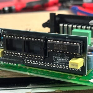 May include: A green circuit board with two black integrated circuits, labelled U1 and U2. The board has a yellow capacitor labelled 105/100 and the text "FLYING BRICK" and "PARTS FOR CLASSIC CARDS".