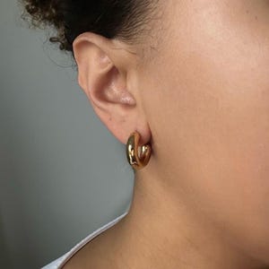 Puede incluir: Pendientes de aro dorados con un dise&ntilde;o liso y redondeado. Los pendientes se llevan en la oreja de una persona.