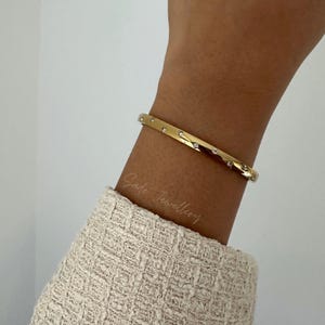 May include: A gold-colored bangle bracelet with small, evenly spaced clear gemstones. The bracelet is worn on a wrist, and the text "Sade Jewellery" is visible on the skin. The bracelet is a simple, elegant piece of jewelry.