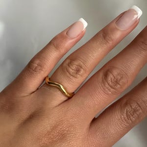 May include: A gold-toned ring with a wavy design. The ring is worn on the middle finger of a hand with white-tipped nails.