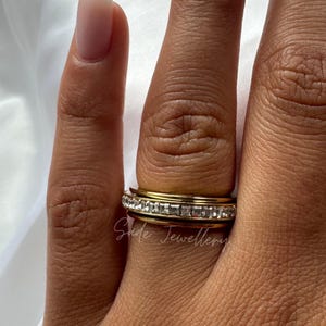 Pode incluir: Close-up de anéis dourados com pedras quadradas transparentes. Os anéis estão empilhados em um dedo. O nome da marca "Sade Jeweller" está escrito em itálico na imagem.