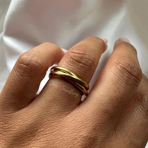 Könnte beinhalten: Ein goldfarbener Ring mit drei ineinander verschlungenen Bändern. Der Ring ist glatt und glänzend und reflektiert das Licht. Der Schmuck wird an einem Finger getragen. Der Hintergrund ist ein weicher, weißer Stoff.