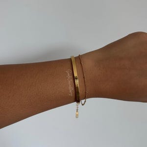 Könnte beinhalten: Ein goldfarbenes Armband-Set an einem Handgelenk. Das Set umfasst einen massiven, flachen Armreif und ein zartes Kettenarmband. Die Armbänder sind schlicht und elegant, mit poliertem Finish. Der Hintergrund ist einfarbig und neutral.
