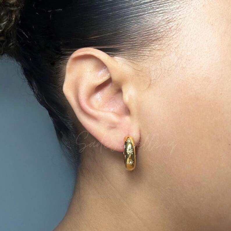 May include: Gold hoop earrings with a starburst design. The earrings are worn in a person's ear.